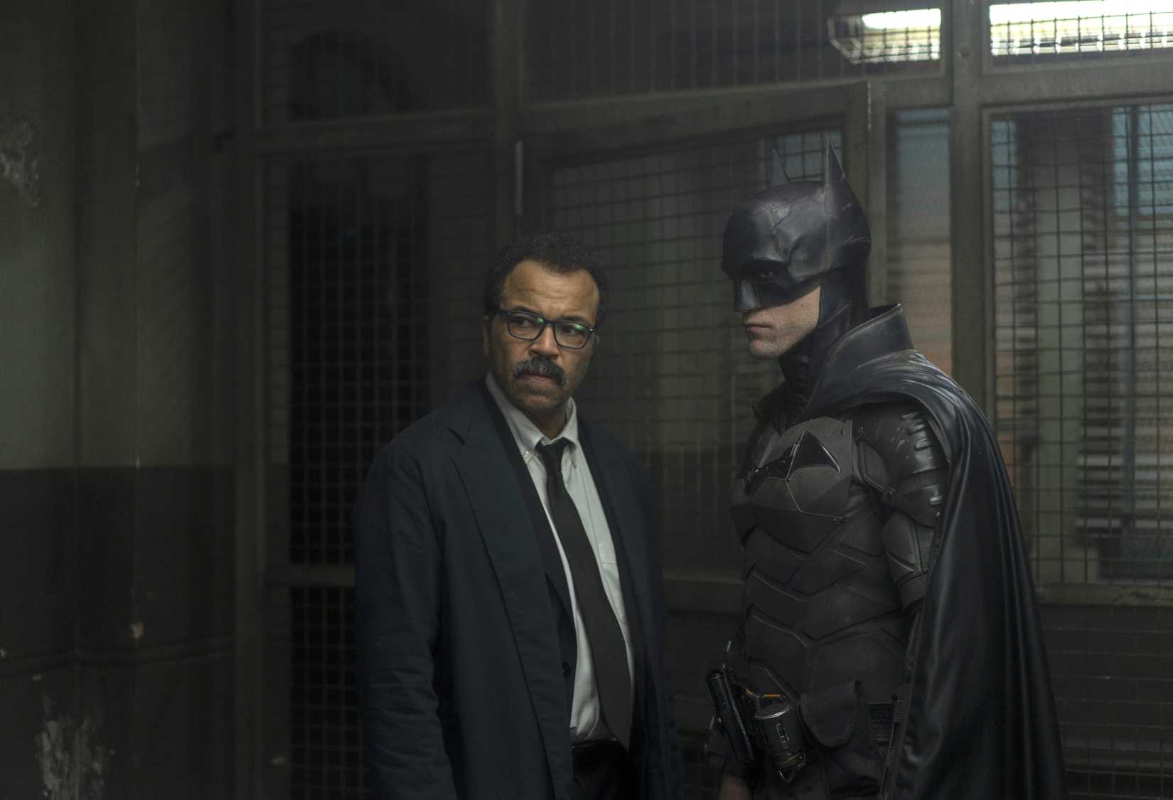 Jeffrey Wright as Lt. James Gordon and Robert Pattinson as Batman in The Batman. They stand facing each other but faces turned out sternly inside a police precinct.