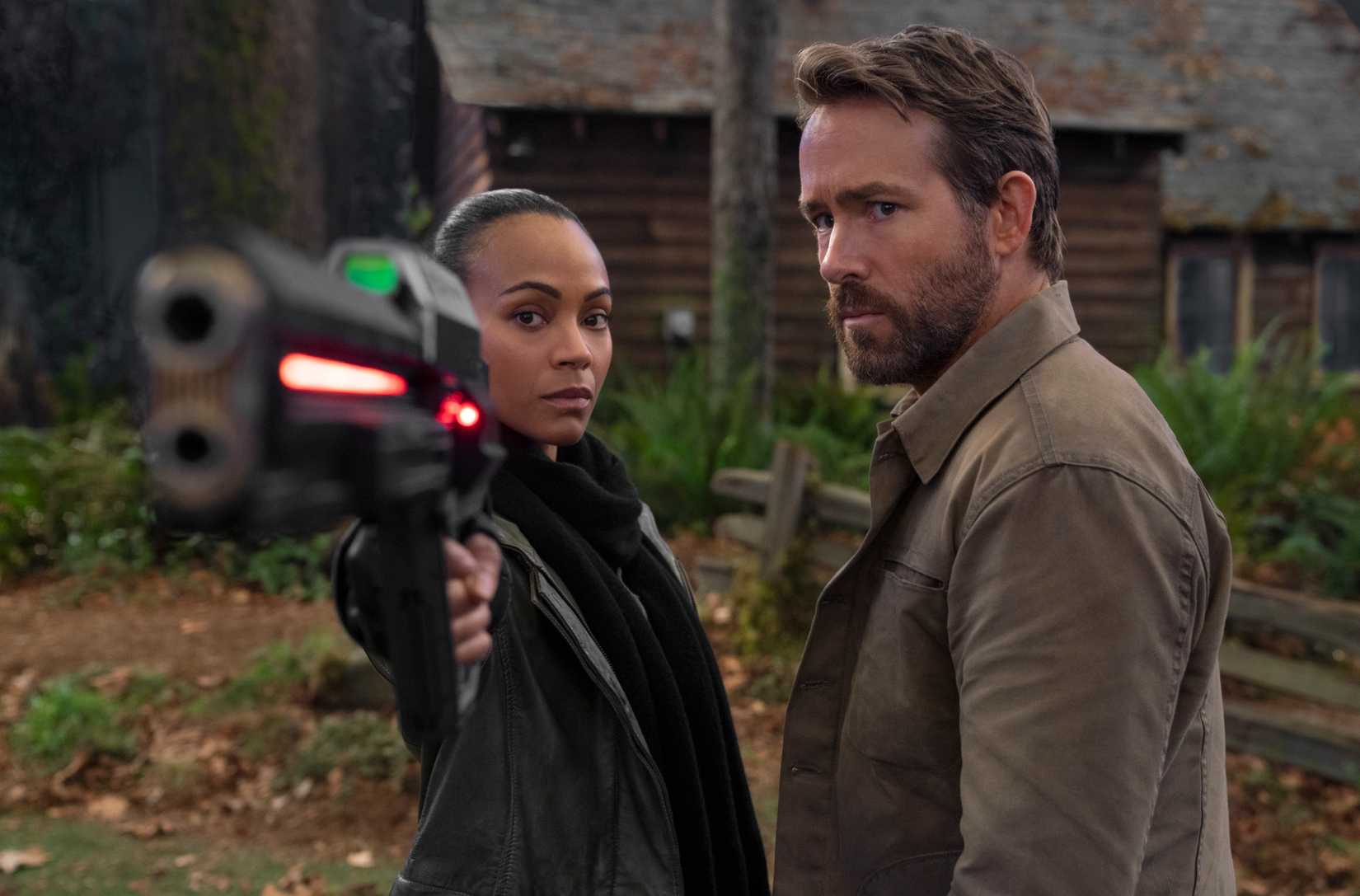 Zoe Saldana and Ryan Reynolds stand side by side outdoors in The Adam Project, with Zoe pointing a giant-ass glowing futuristic gun at the camera