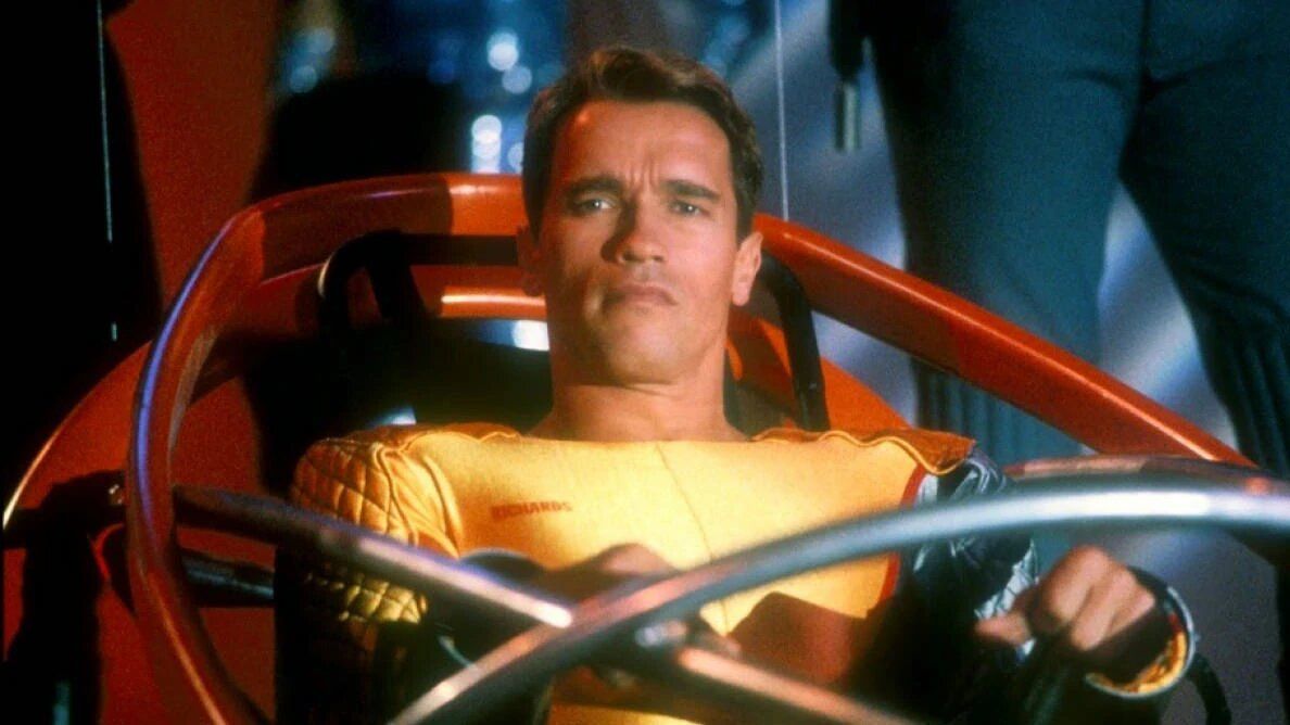 Arnold Schwarzenegger as Ben Richards in a yellow jumpsuit seated in a futuristic seat in The Running Man.