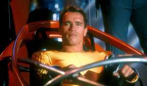 Arnold Schwarzenegger as Ben Richards in a yellow jumpsuit seated in a futuristic seat in The Running Man.