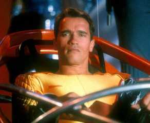 Arnold Schwarzenegger as Ben Richards in a yellow jumpsuit seated in a futuristic seat in The Running Man.