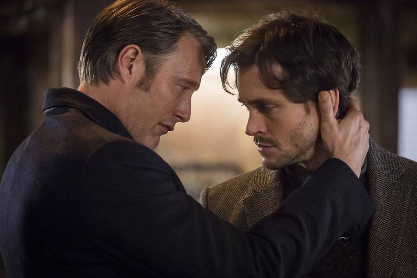 Mads Mikkelsen as Hannibal Lecter stands close to Hugh Dancy as Will Graham in Hannibal. Their foreheads are almost touching, Hannibal has his hand on the back of Will's neck, and they both look solemn.