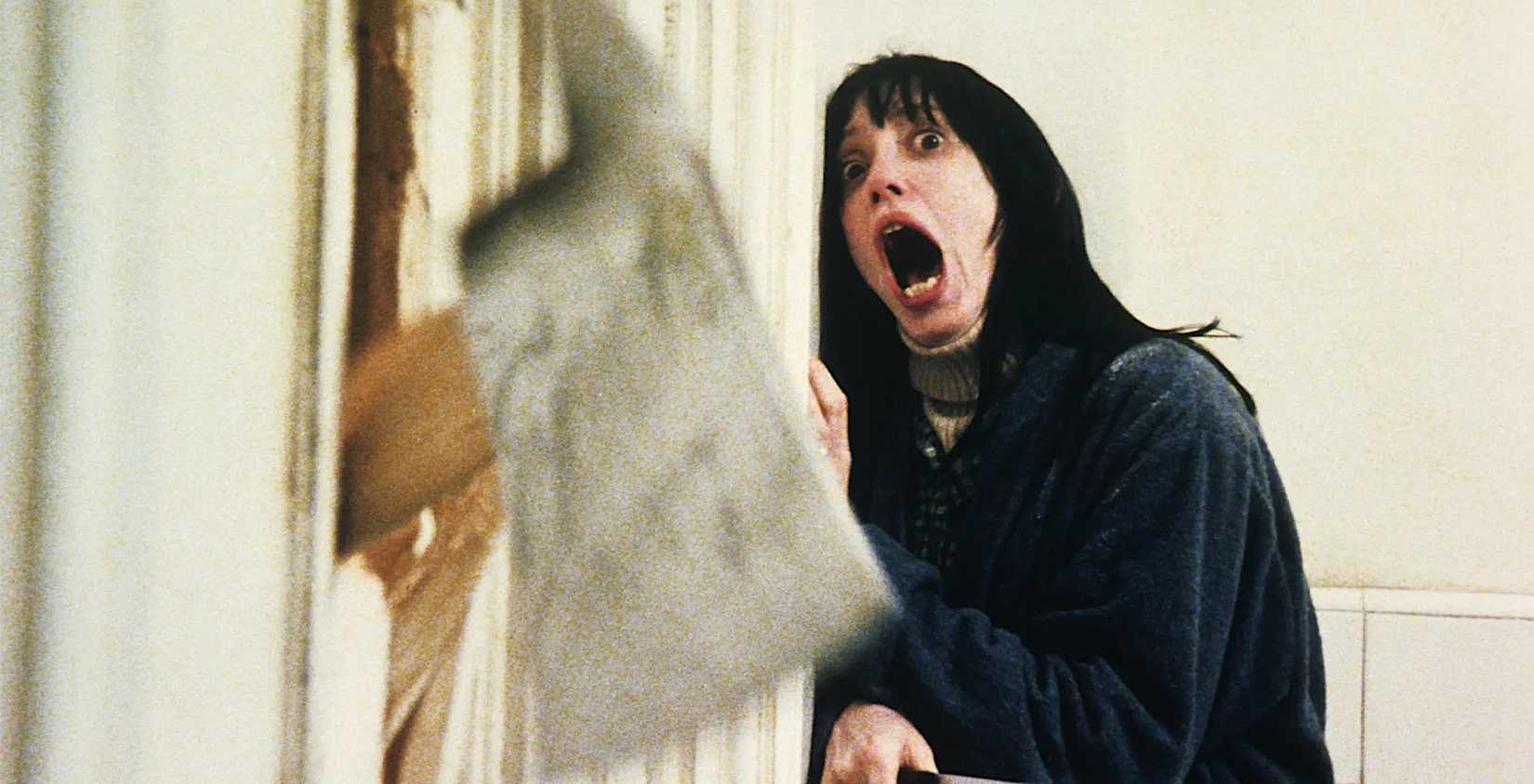 Shelley Duvall screams in terror and presses her body into a corner as an axe crashes through the door of the bathroom she’s hiding in in Stanley Kubrick’s The Shining