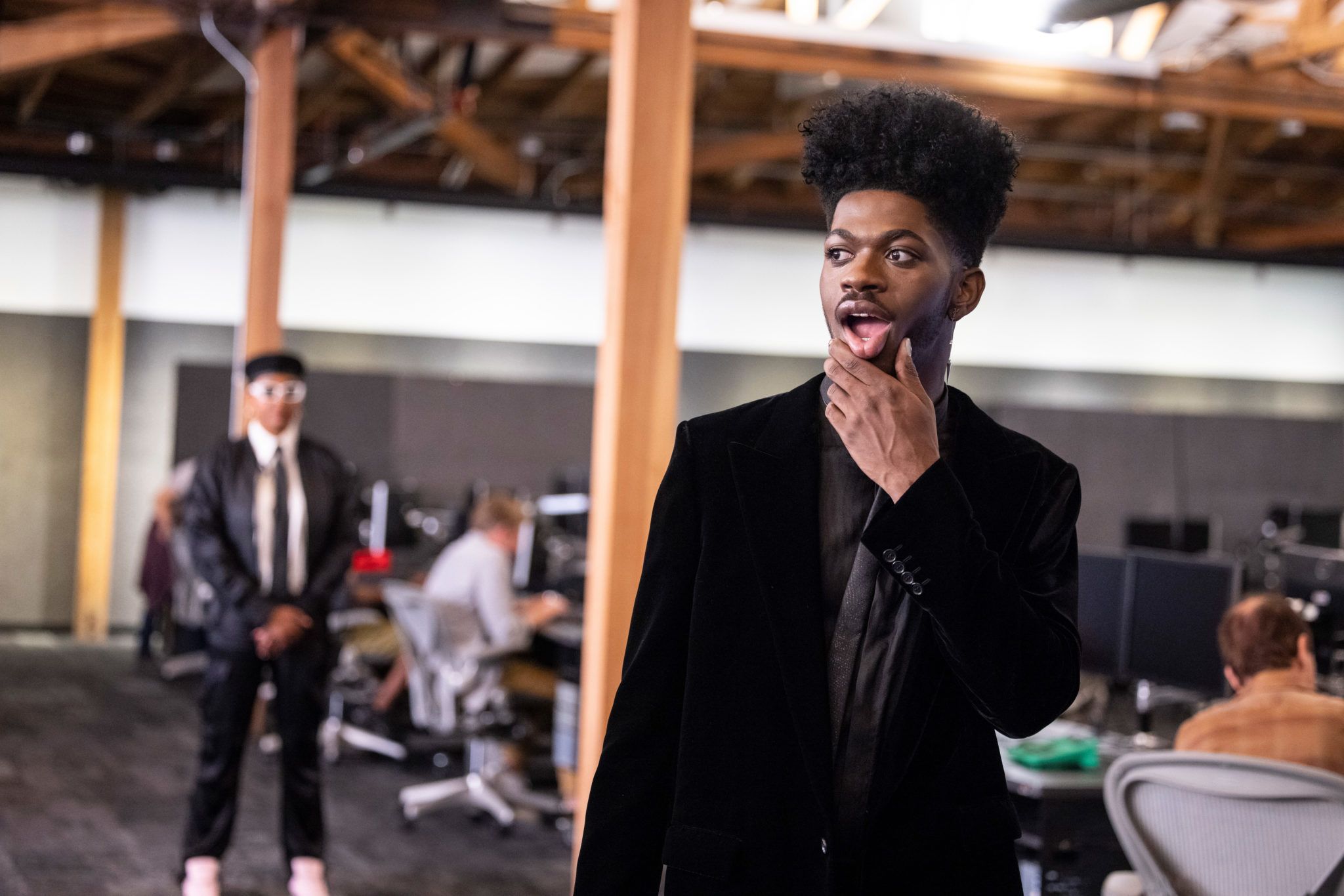 Lil Nas X, a famous pop star and young Black man, stands in the work area of Riot HQ. He is wearing a suit and looks around his surroundings, running a thumb along the side of his mouth. Another employee is in the background, out of focus.