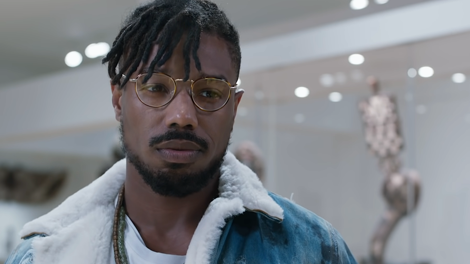 Erik Killmonger (Michael B. Jordan) in a fleece-lined jean jacket and glasses stands in a museum in 2018’s Black Panther