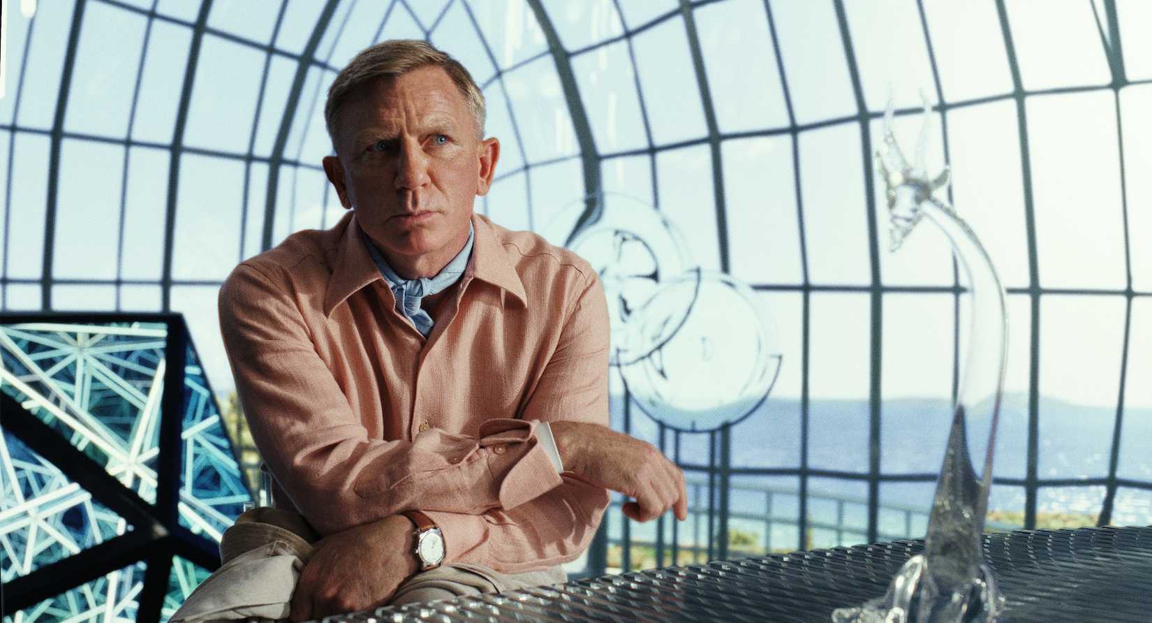 Daniel Craig รับบทเป็น Benoit Blanc ใน Glass Onion ใส่ใจในเสื้อเชิ้ตผ้าลินินสีชมพูและเน็คไทสีน้ำเงินในโดมแก้วหลายรูปทรง
