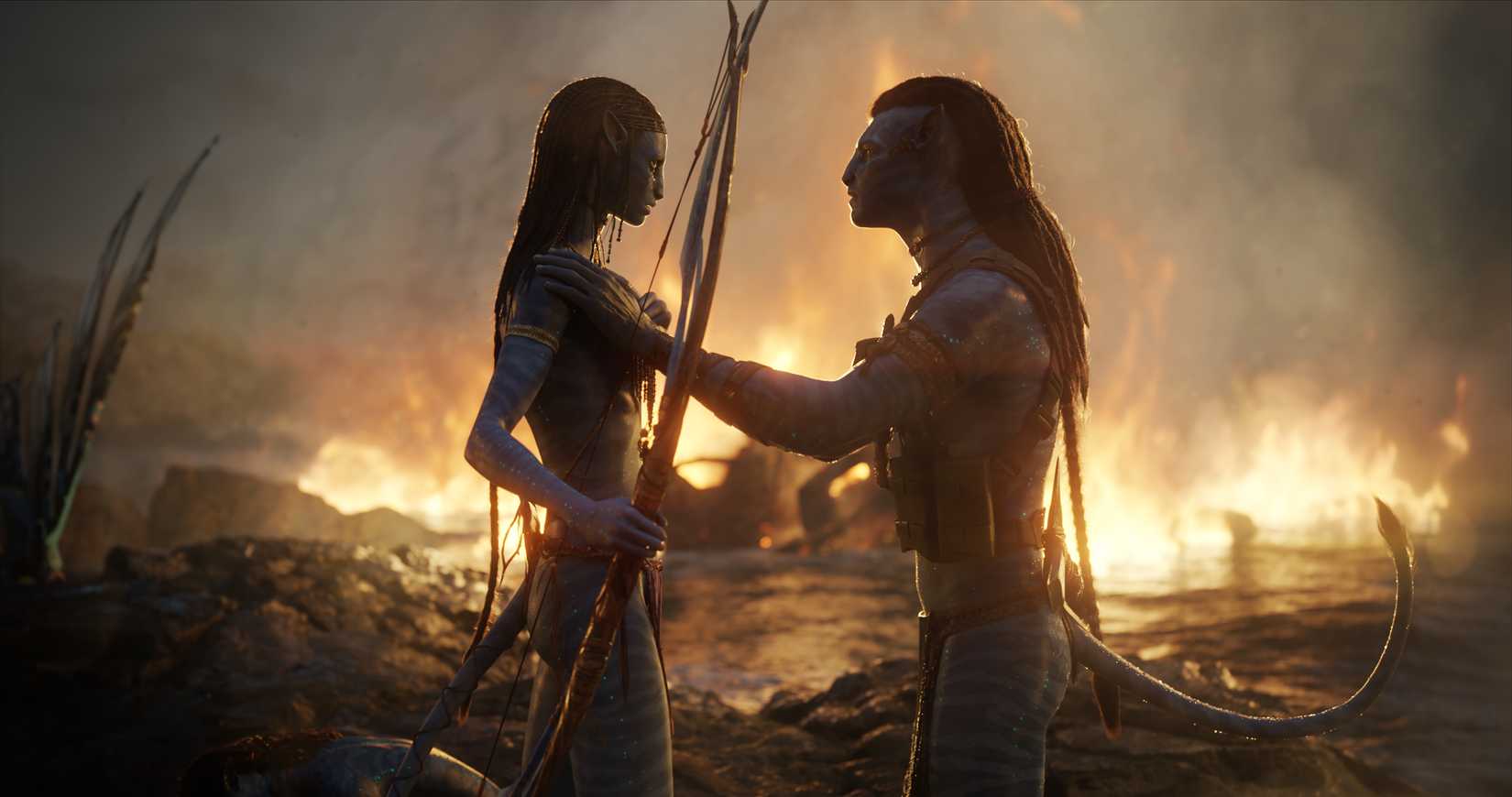 Jake Sully and Neytiri comfort each other after battle, with a fire roaring behind them over the water in Avatar: The Way of Water