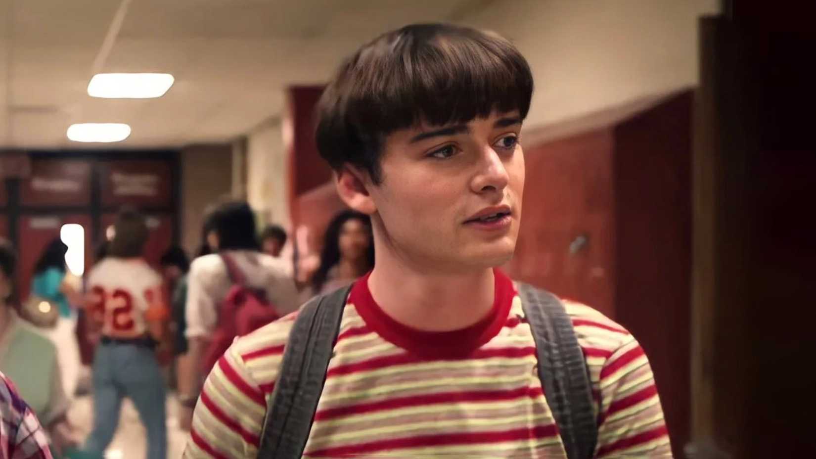 Noah Schnapp as Will Byers, walking through the hall of high school with a bowl cut and a striped red and yellow shirt while wearing a backpack in Stranger Things