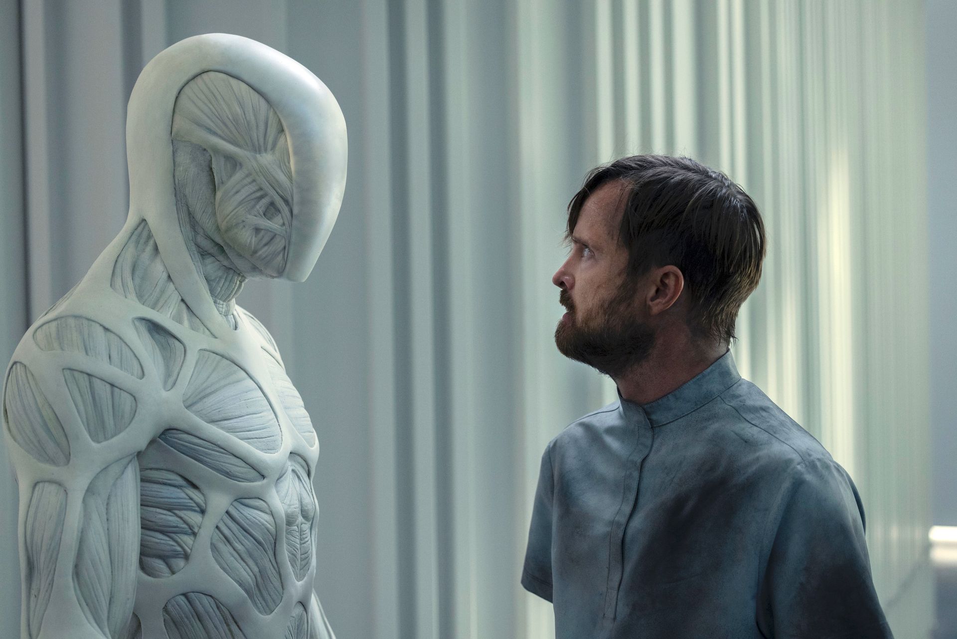 A spindly muscular white robot stands face to face with a white bearded man in a hospital gown in Westworld season 4