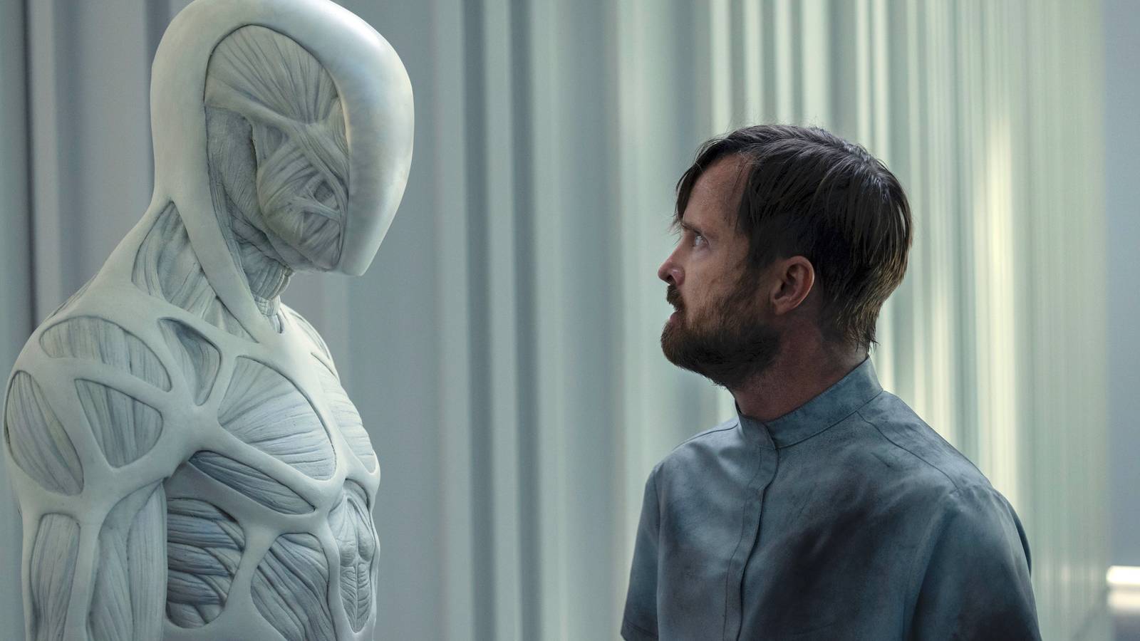 A spindly muscular white robot stands face to face with a white bearded man in a hospital gown in Westworld season 4