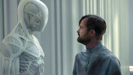 A spindly muscular white robot stands face to face with a white bearded man in a hospital gown in Westworld season 4