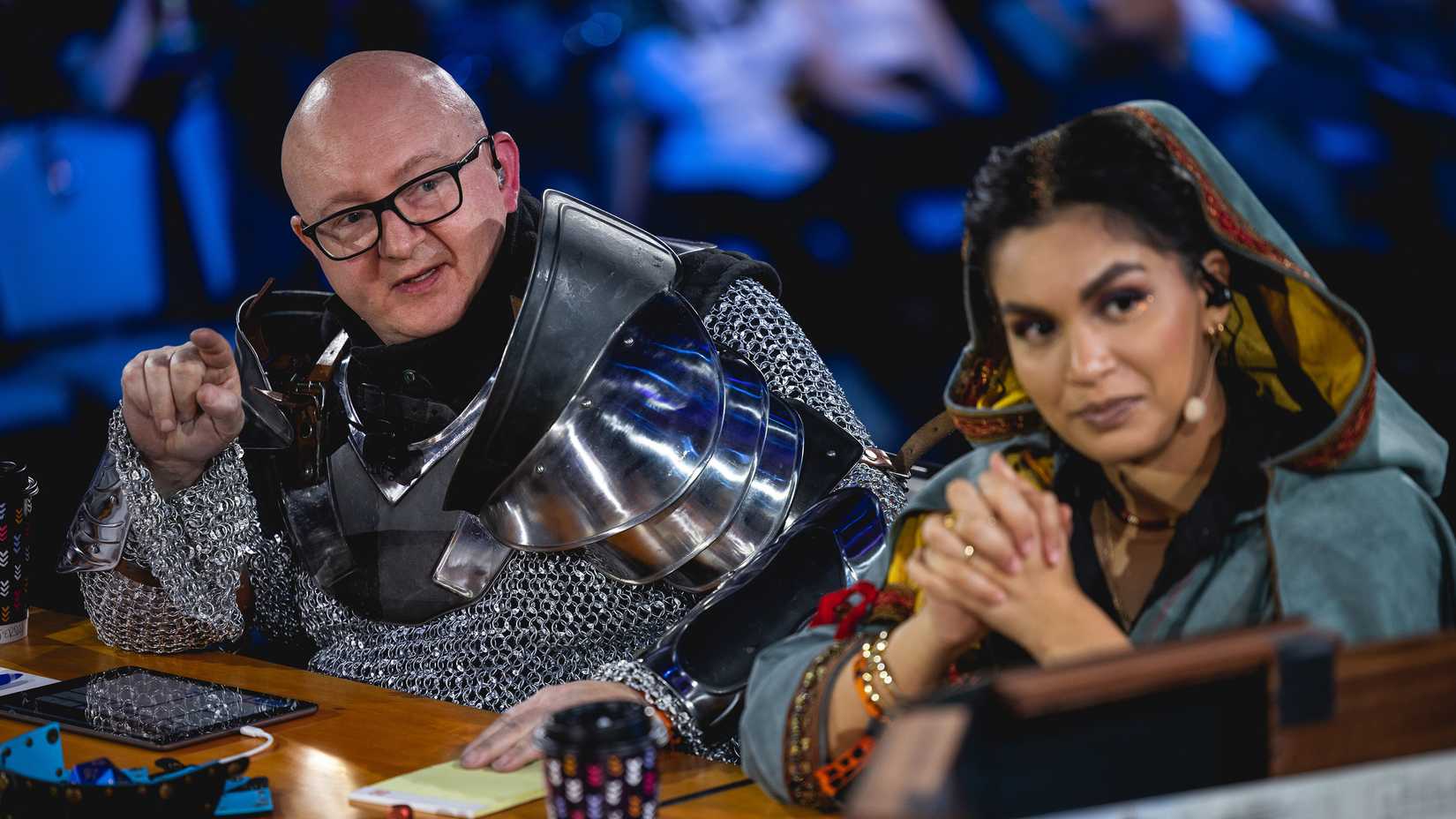 Jerry Holkins wearing chainmail and armor sits next to Jasmine Bhullar in a staged performance of Acquisitions Incorporated.
