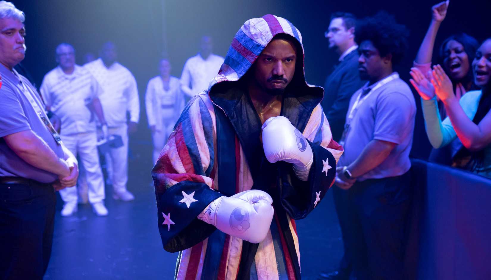 Adonis Creed (Michael B. Jordan) se prepara para entrar en un ring de boxeo, con guantes blancos y una túnica con capucha temática de bandera americana roja, blanca y azul en Creed III