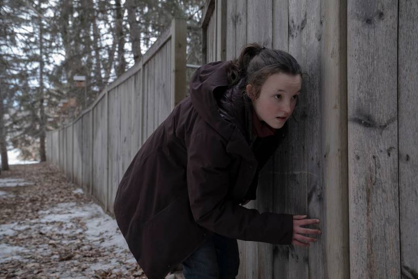 Ellie crouches and presses up against a wooden fence in winter in a scene from HBO’s The Last of Us