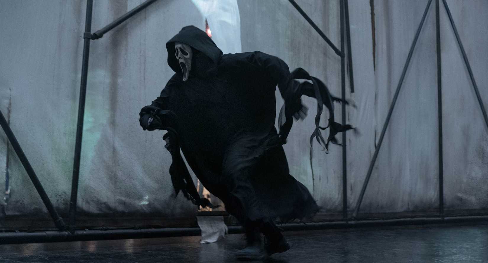 Ghostface chasing a victim through a high-contrast set with white curtains and scaffolding in Scream VI