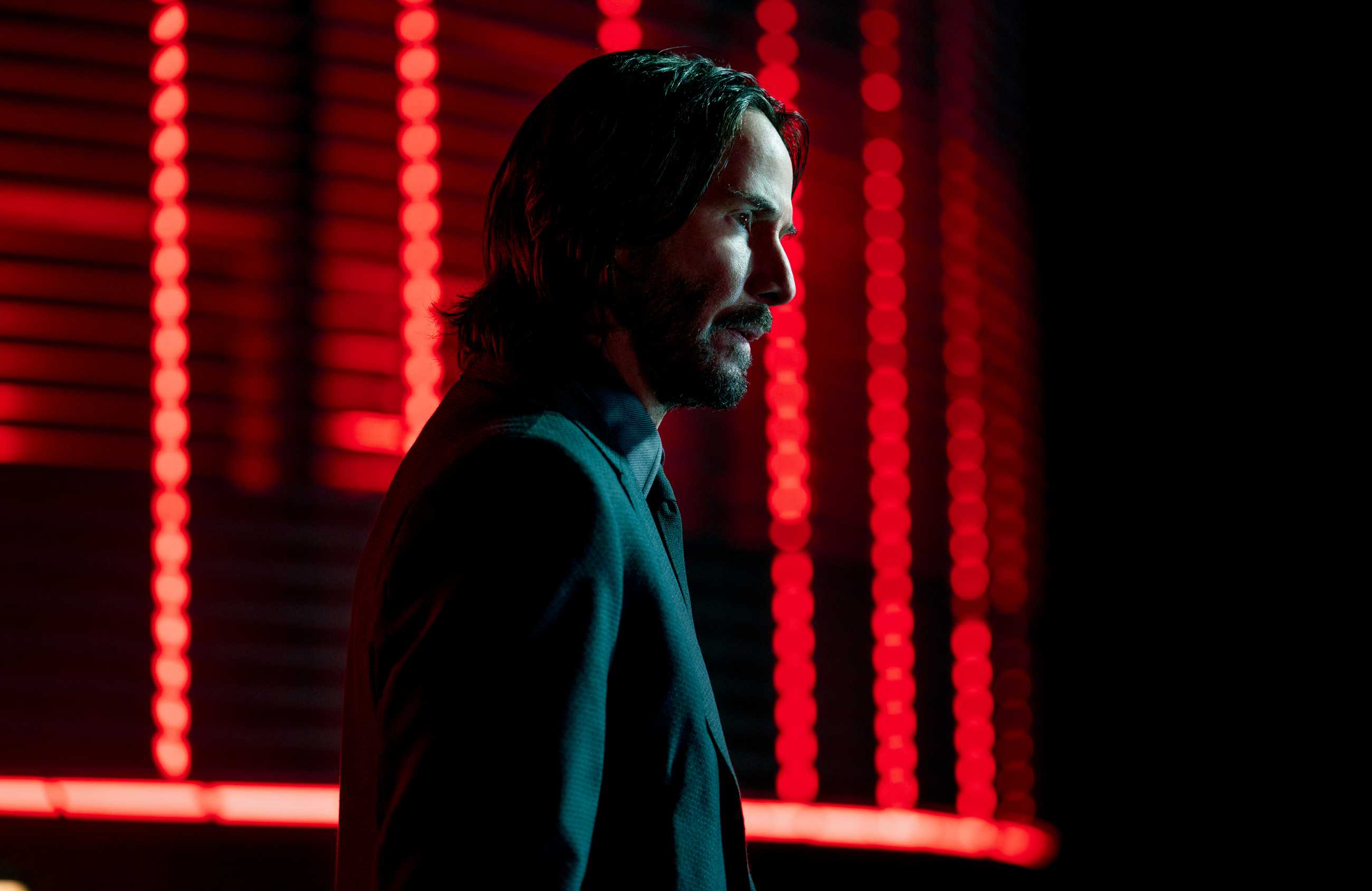 Keanu Reeves as John Wick, standing in profile in front of red lights.