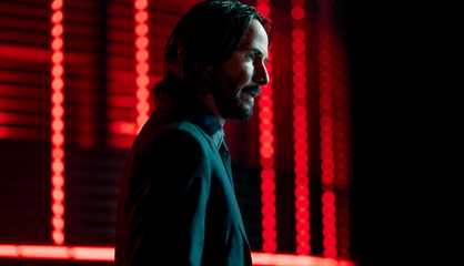 Keanu Reeves as John Wick, standing in profile in front of red lights.