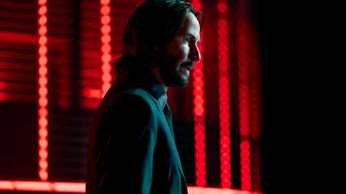 Keanu Reeves as John Wick, standing in profile in front of red lights.