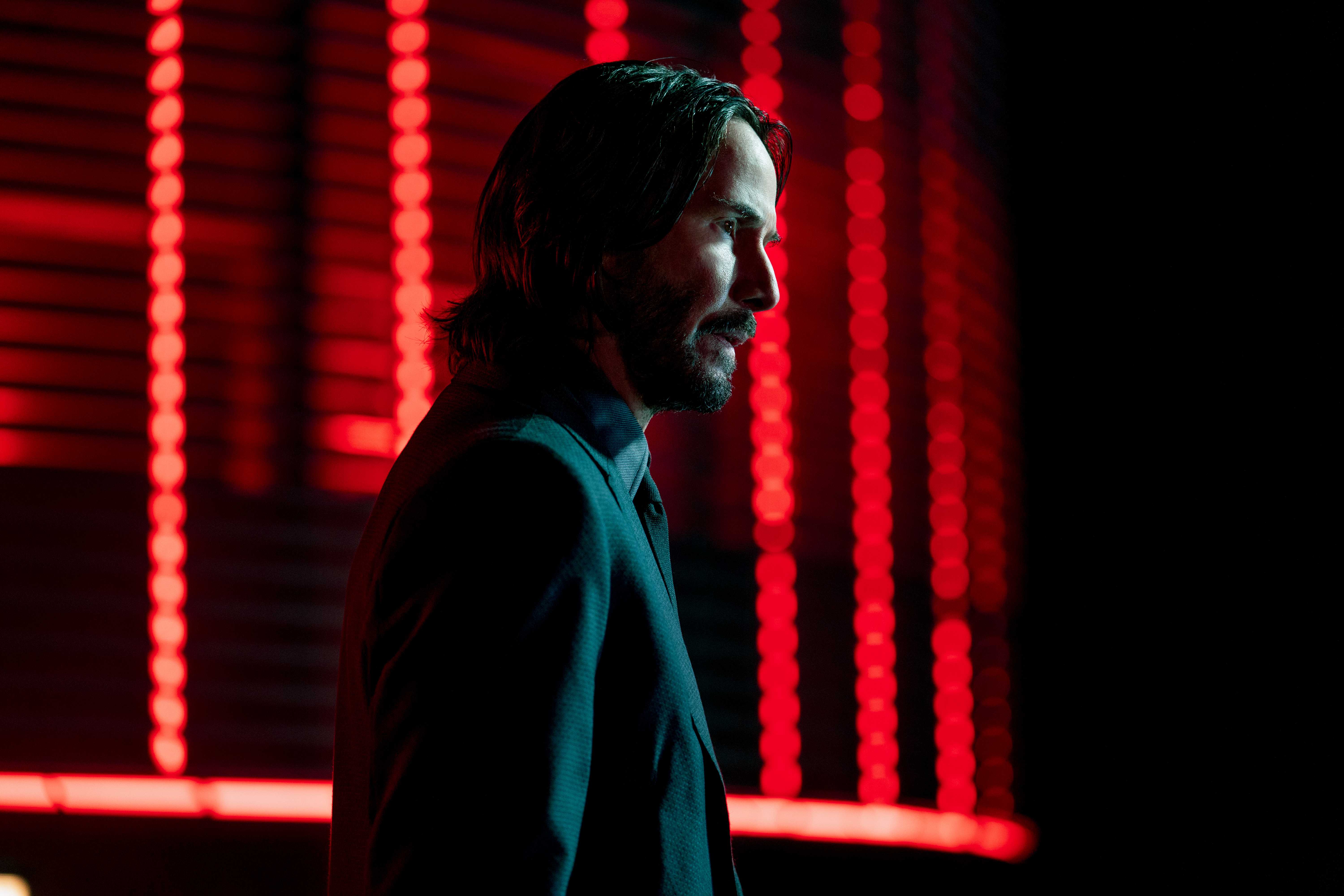Keanu Reeves as John Wick, standing in profile in front of red lights.