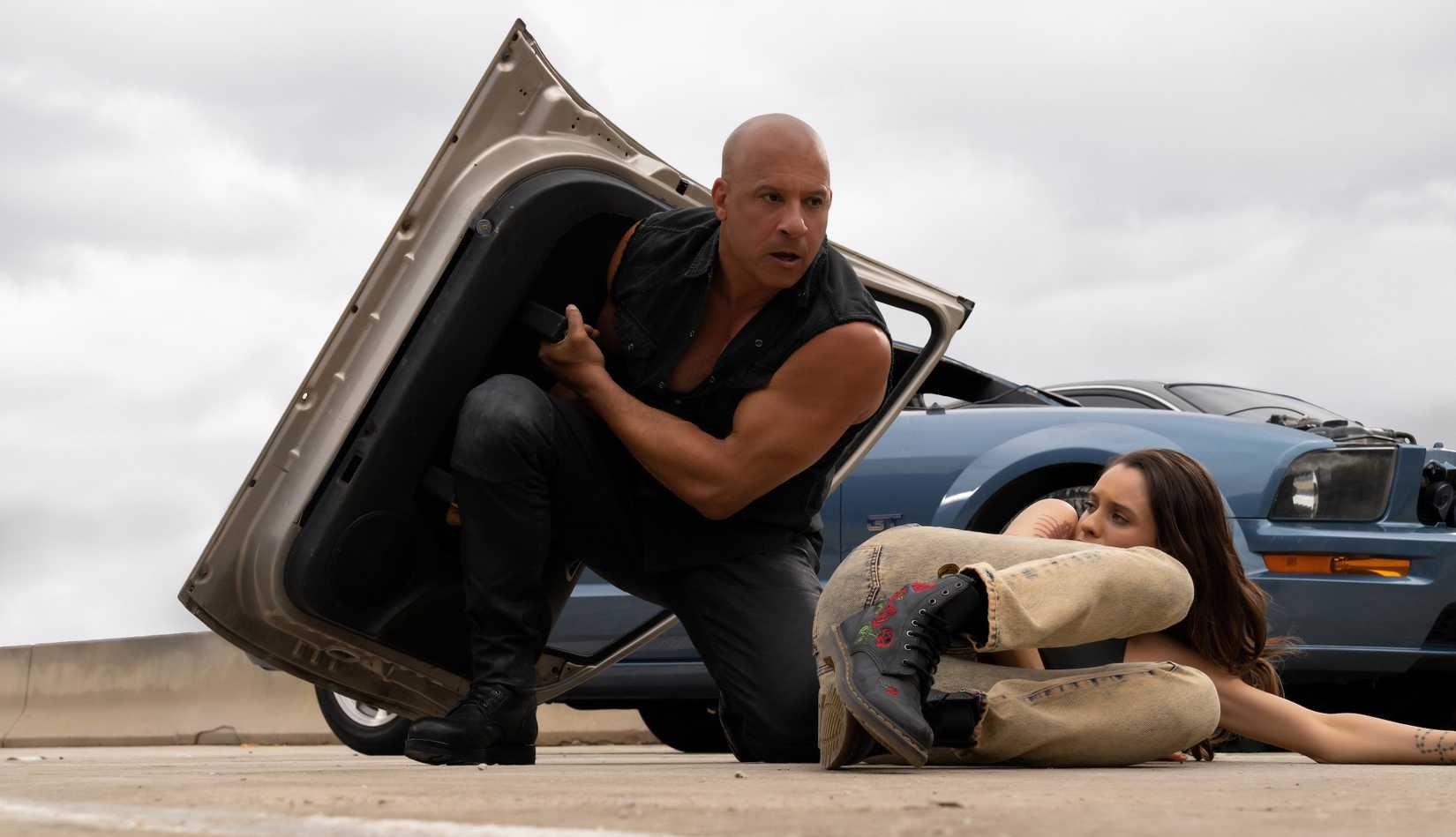 Dom Toretto (Vin Diesel) holds up a detached car door to shield himself from a hail of bullets as he moves to cover Isabel (Daniela Melchior), lying on the ground in front of a car, in Fast X