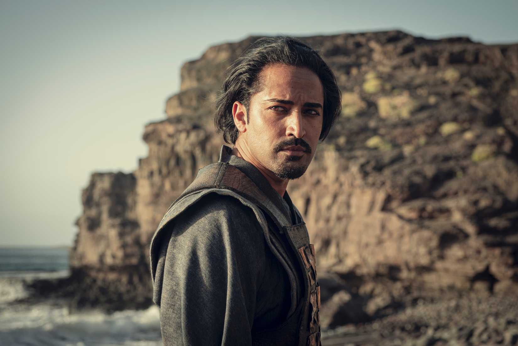 Vilgefortz (Mahesh Jadu) stands tall on a rocky beach looking pensively to his right in The Witcher.