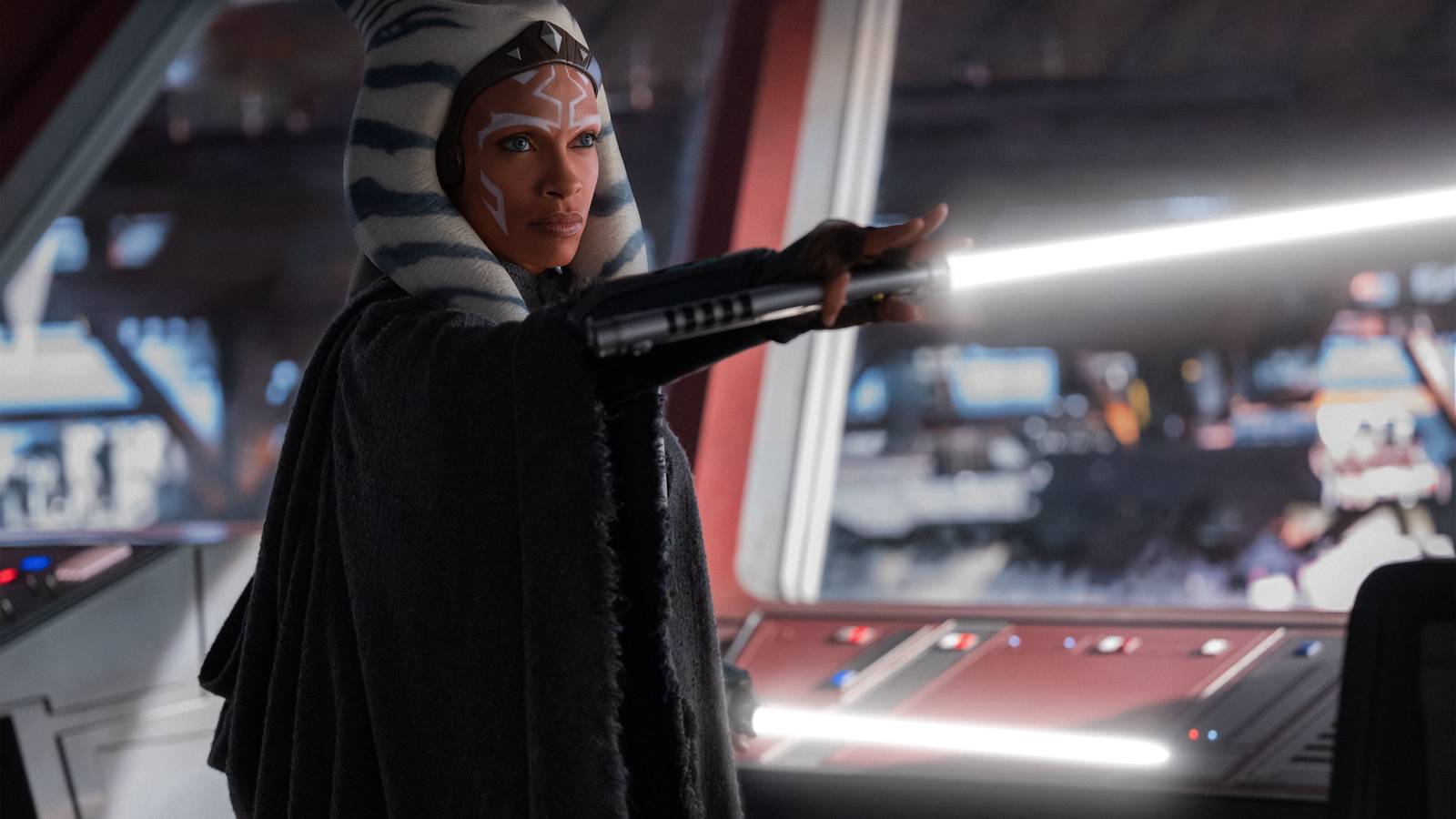Ahsoka stands in front of a window in a cloak holding here white lightsaber horizontally in front of her in a scene from the Disney Plus series Ahsoka