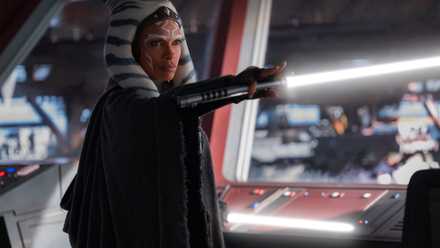 Ahsoka stands in front of a window in a cloak holding here white lightsaber horizontally in front of her in a scene from the Disney Plus series Ahsoka