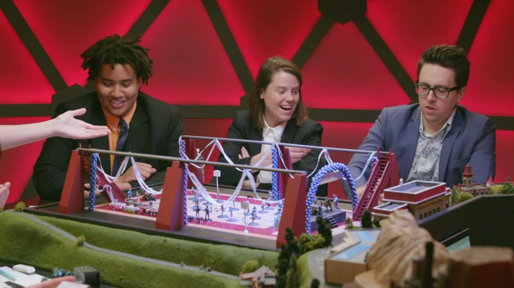 Lou Wilson, Ally Beardsley, and Brian Murphy during an episode of Fantasy High. There’s a big dance spread out in miniature at the table.