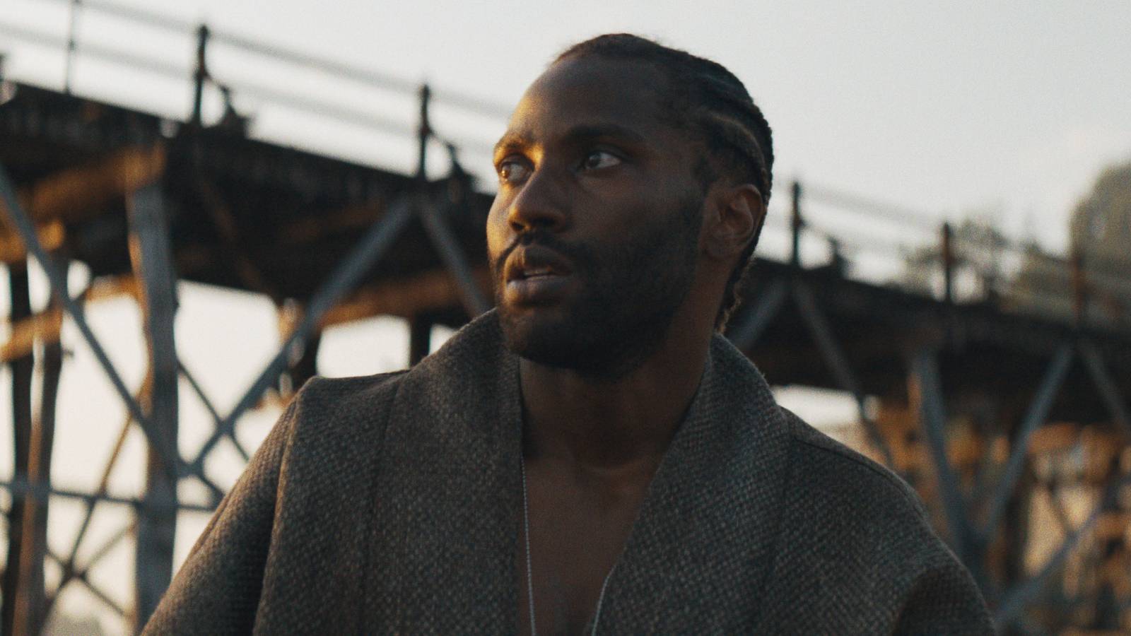 John David Washington as Joshua, looking up at something off camera with a wooden bridge behind him at sunset in The Creator