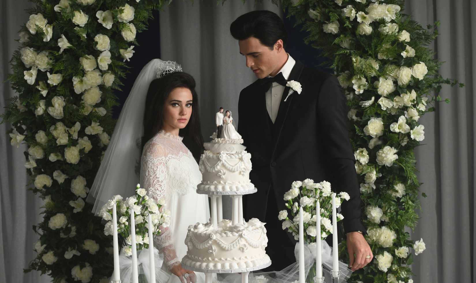 Priscilla Presley (Cailee Spaeny, in a white wedding dress and swept-back white veil), stands with Elvis Presley (Jacob Elordi, in a black tuxedo) stand behind their tiered white wedding cake under an arch of green leaves and white flowers, with Elvis looking downward and Priscilla looking directly into the camera, in Sofia Coppola’s Priscilla