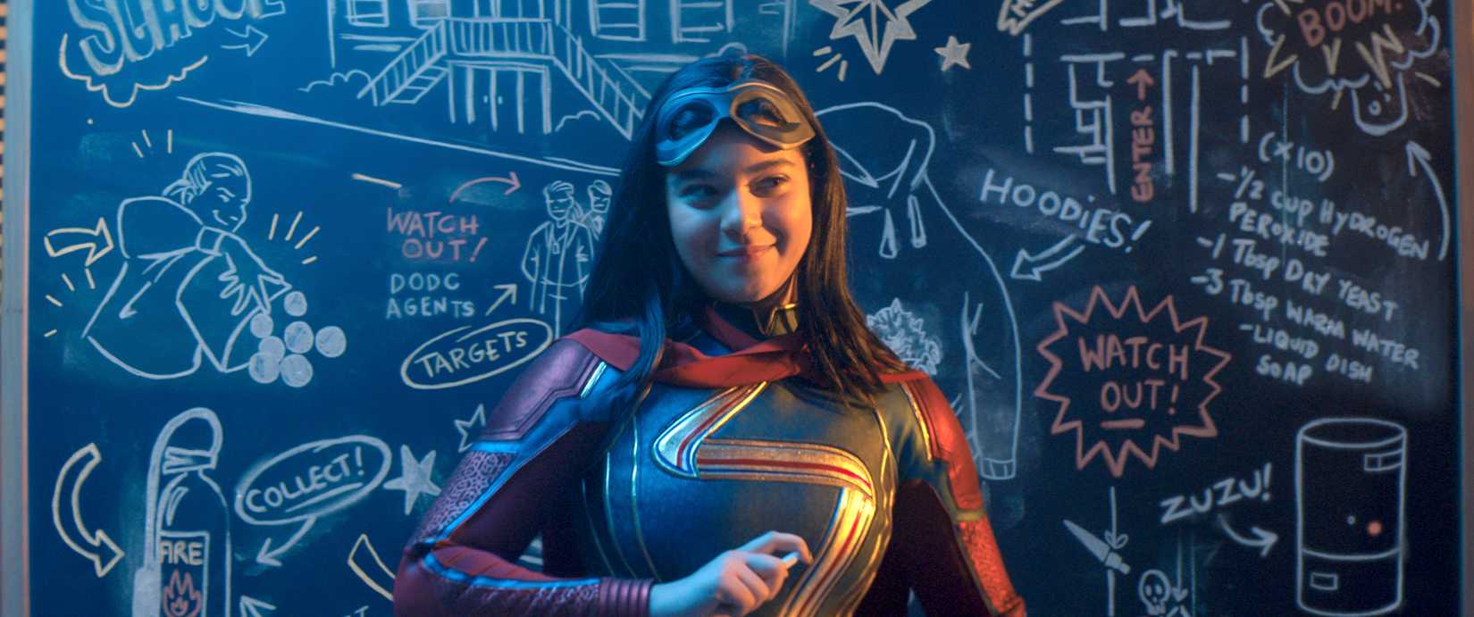 Kamala Khan/Ms. Marvel stands in front of a crowded chalkboard, smirking and holding a piece of chalk. Her mask is pushed up onto her forehead. 