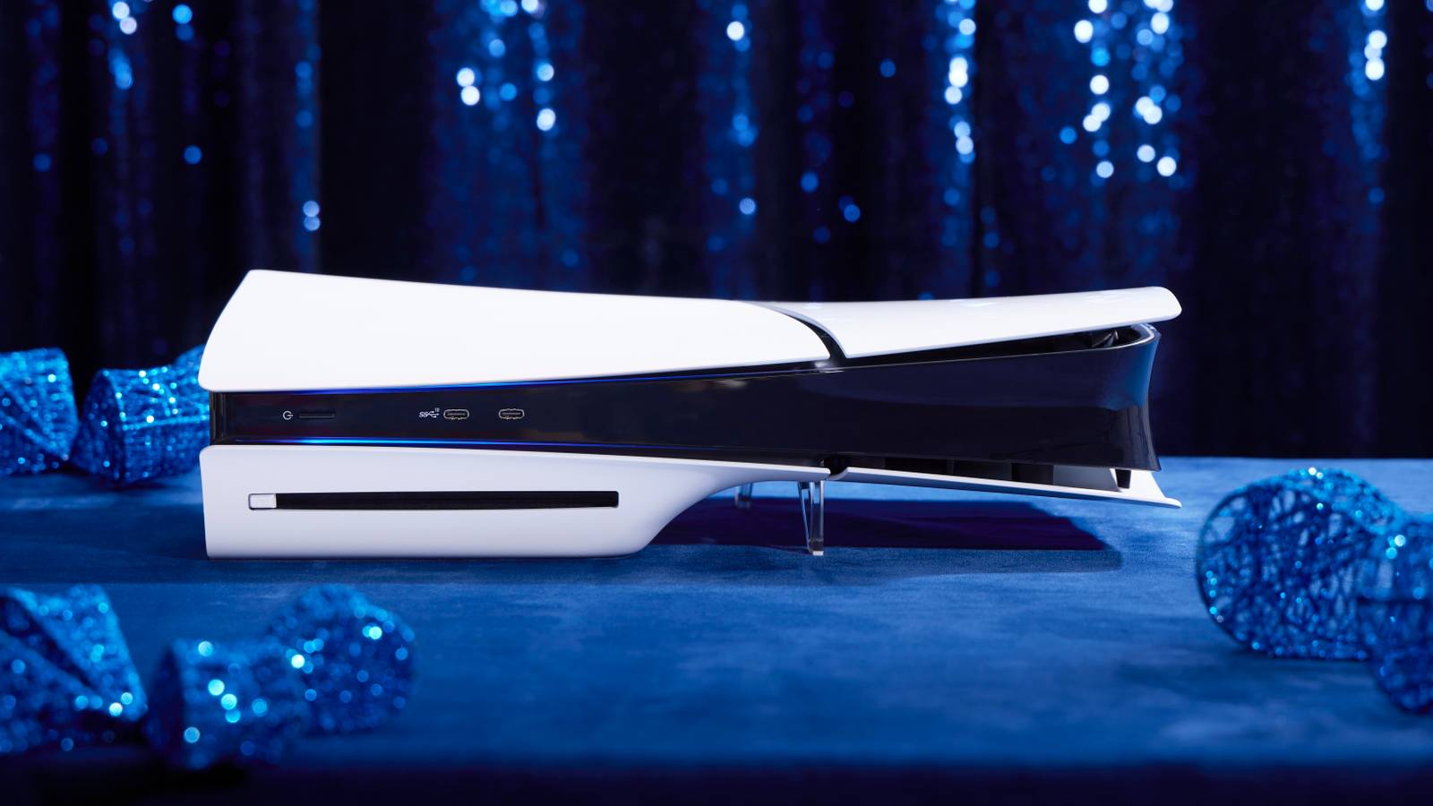 A photograph of Sony’s slim PS5 console laying horizontally on a blue suede surface. There are blue holiday ornaments surrounding the console that are slightly out of focus, as well as a sparkling blue curtain behind the console.
