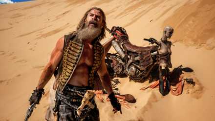 Chris Hemsworth standing in the desert in front of a motorcycle with his shirt open and a weapon in his hand in Furiosa