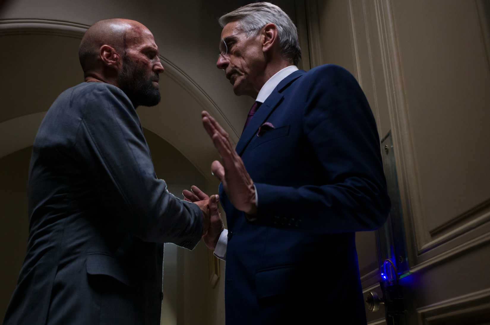 Jason Statham bullies Jeremy Irons, who raises his hands up in defense while blocking a door, in The Beekeeper