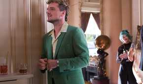 Josh Hutcherson, wearing a green suit and looking like the kind of guy you definitely do not want to take home to meet your parents, smiles by some drinks in The Beekeeper