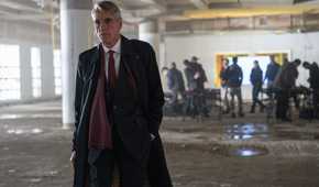 Jeremy Irons, wearing a long coat, walks towards the camera and away from uniformed people in what looks like a parking garage in The Beekeeper.