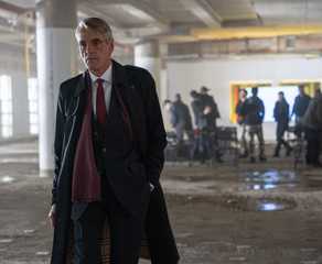 Jeremy Irons, wearing a long coat, walks towards the camera and away from uniformed people in what looks like a parking garage in The Beekeeper.
