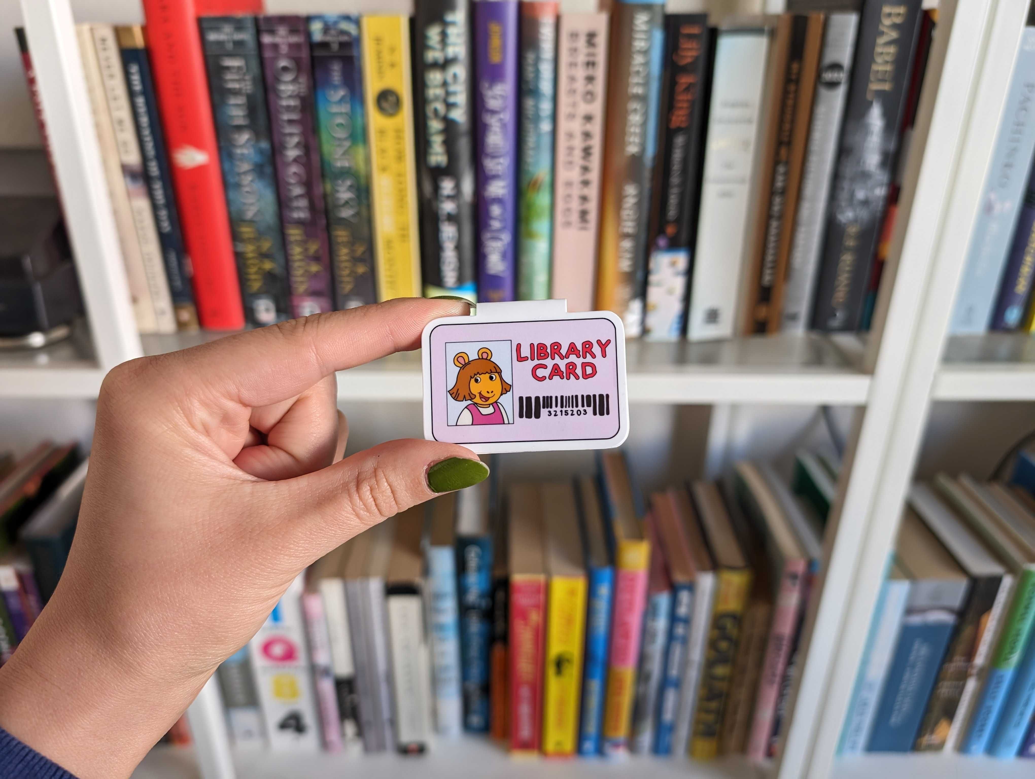 A small D.W. library card, from Arthur, held in front of a book shelf. The hand is light-skinned, with green nail polish.