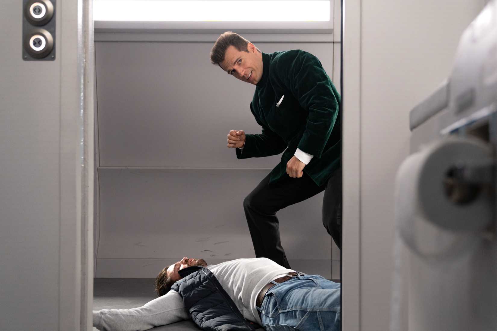 Henry Cavill, with a flat top haircut and green velvet suit, poses over a knocked-out foe in a train carriage in Argylle