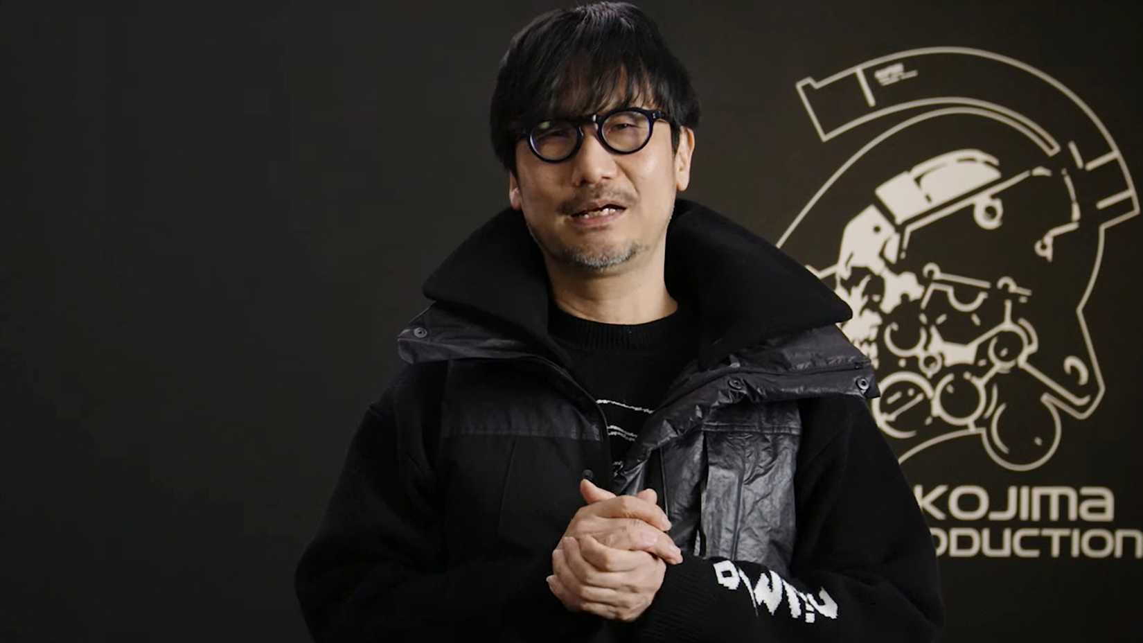 Hideo Kojima stands in front of the Kojima Productions logo in a video promoting the game PHYSINT