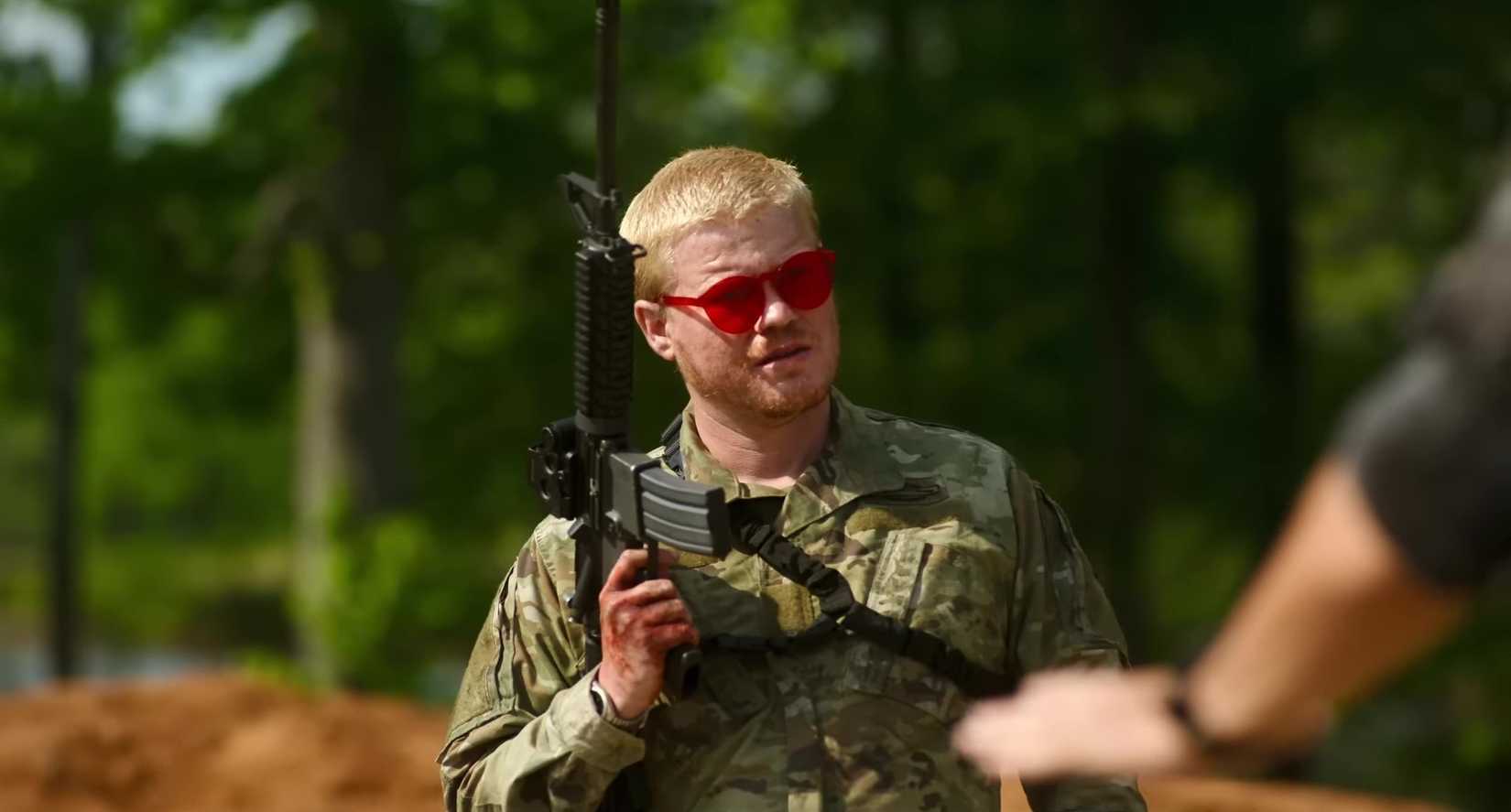 Jesse Plemons trong trang phục quân đội, đeo kính râm đỏ và mang theo súng trường
