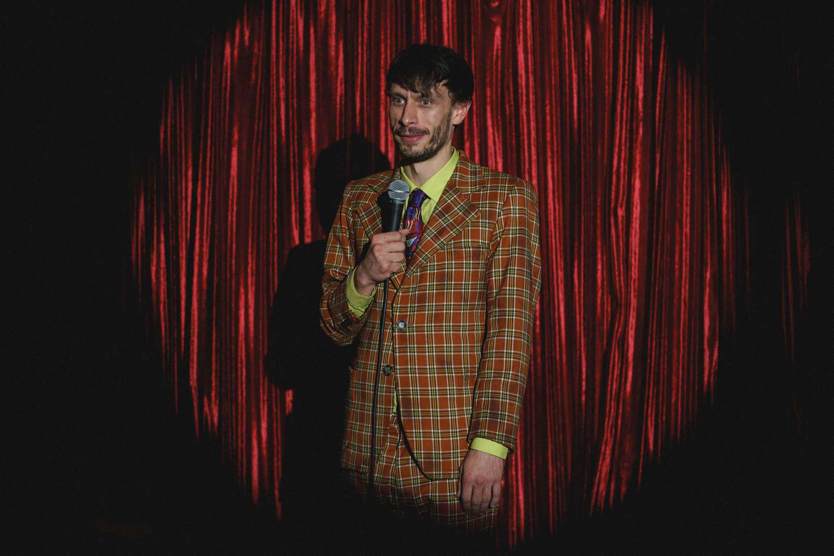Richard Gadd in Baby Reindeer, standing in a loud suit with a microphone in a spotlight in front of a red curtain