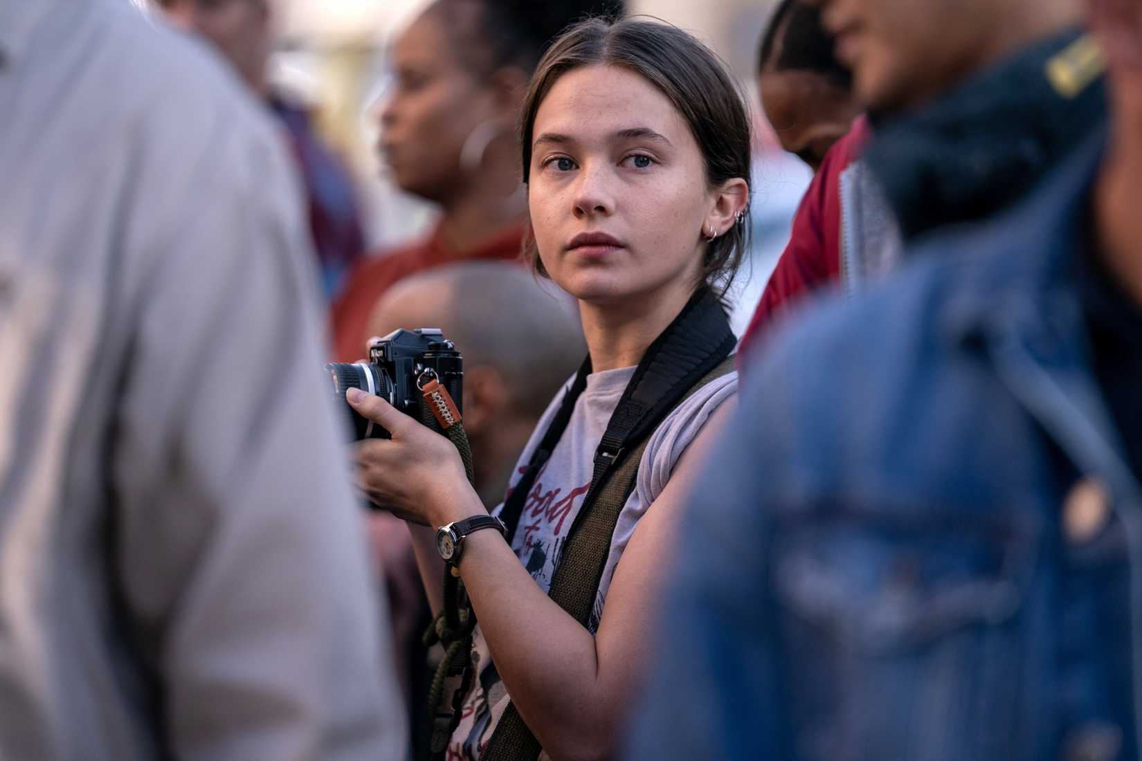 Jessie (Cailee Spaeny), a young photojournalist holding a camera, stands in the middle of a crowd in Alex Garland’s Civil War