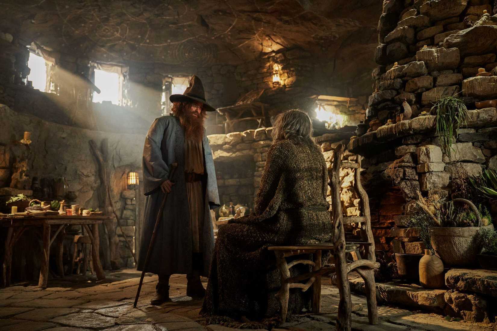 Tom Bombadil stands over a seated Stranger in The Lord of the Rings: The Rings of Power. They’re in a rustic looking cabin. 