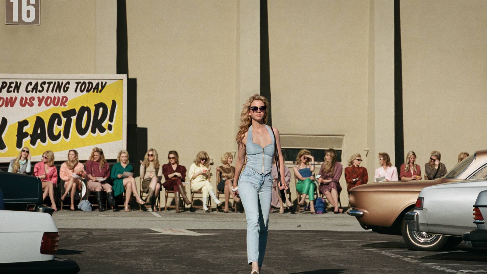 Maxine (Mia Goth) struts across a Hollywood parking lot outside a movie soundstage, with a row of other auditioners lined up in chairs behind her, in Maxxxine 