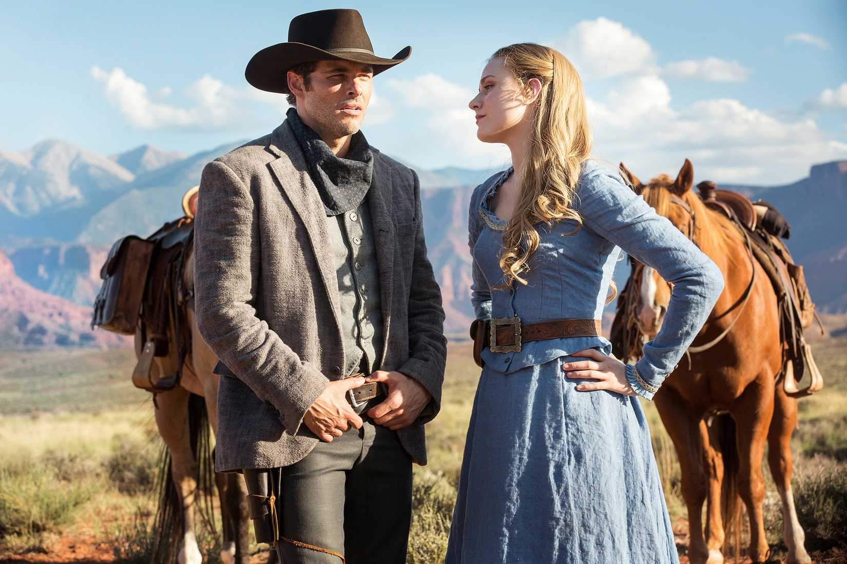 Teddy and Dolores, a man in a cowboy hat and a woman in a sky-blue Old West dress, stand in front of horses on a prairie in Westworld