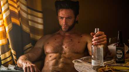 Wolverine, played by Hugh Jackman, sits in a chair. His expression is clearly irritated as he sits shirtless, one hand gripping a bottle of what’s probably alcohol.